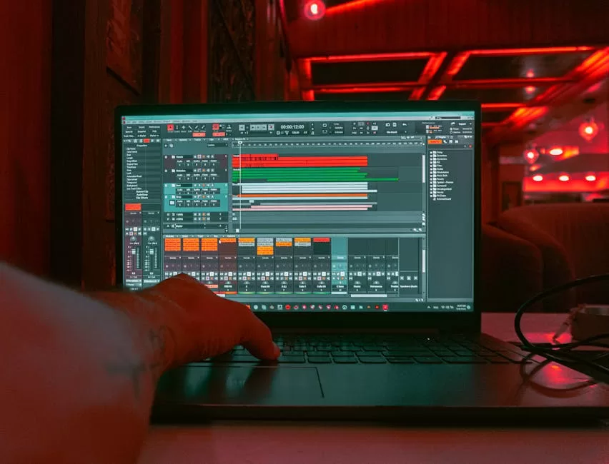 A person editing audio tracks on a laptop indoors with a red ambient lighting background.